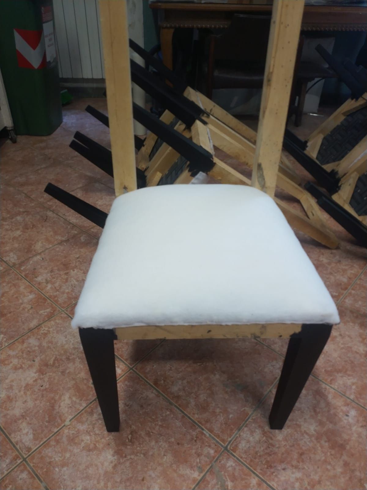 Una sedia di legno con seduta appena rivestita in tessuto bianco, posizionata su un pavimento piastrellato, con altre sedie nelle vicinanze.