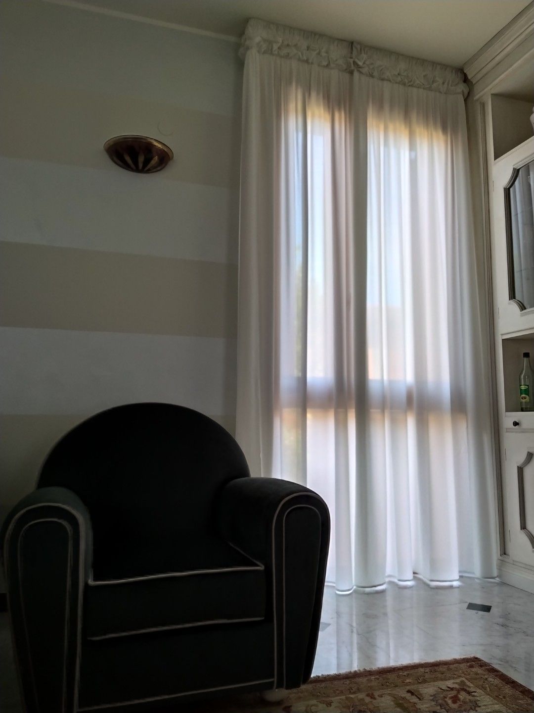 Una poltrona nera è posizionata in un angolo accanto a una finestra con tende bianche lunghe fino al pavimento, vicino a una parete a righe beige e bianche.