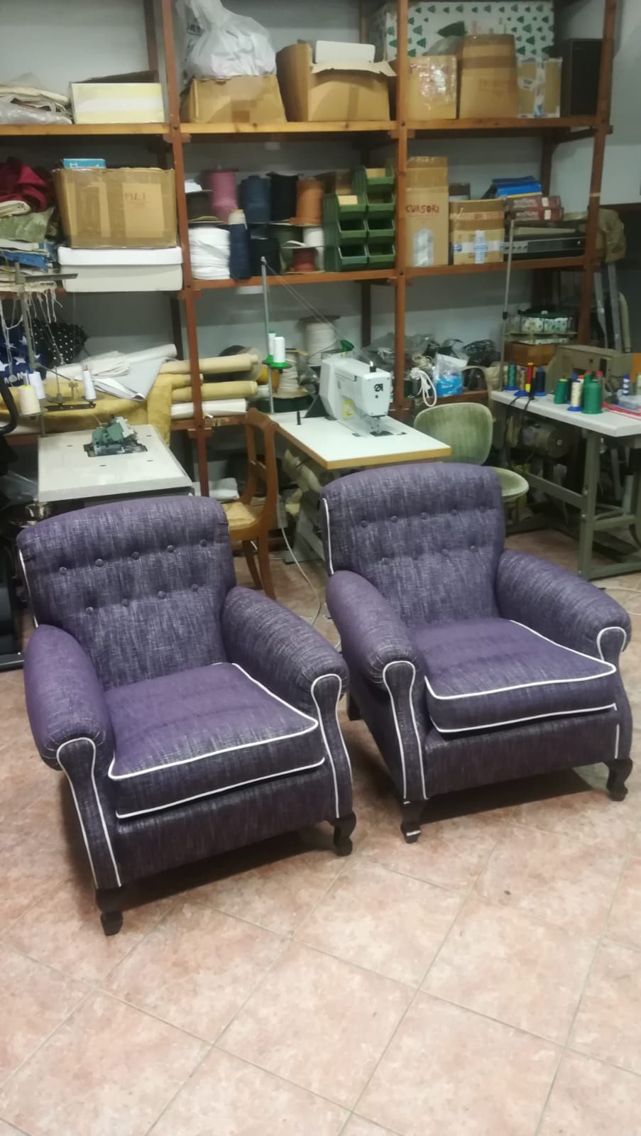 In un laboratorio di cucito o di tappezzeria disordinato, due poltrone imbottite di colore viola con profili bianchi sono disposte una accanto all'altra.
