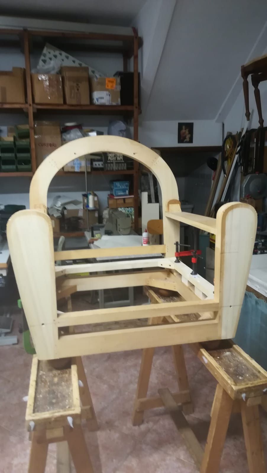 In un'officina, la struttura in legno di una sedia poggia su due cavalletti, mostrando la costruzione essenziale di una poltrona.