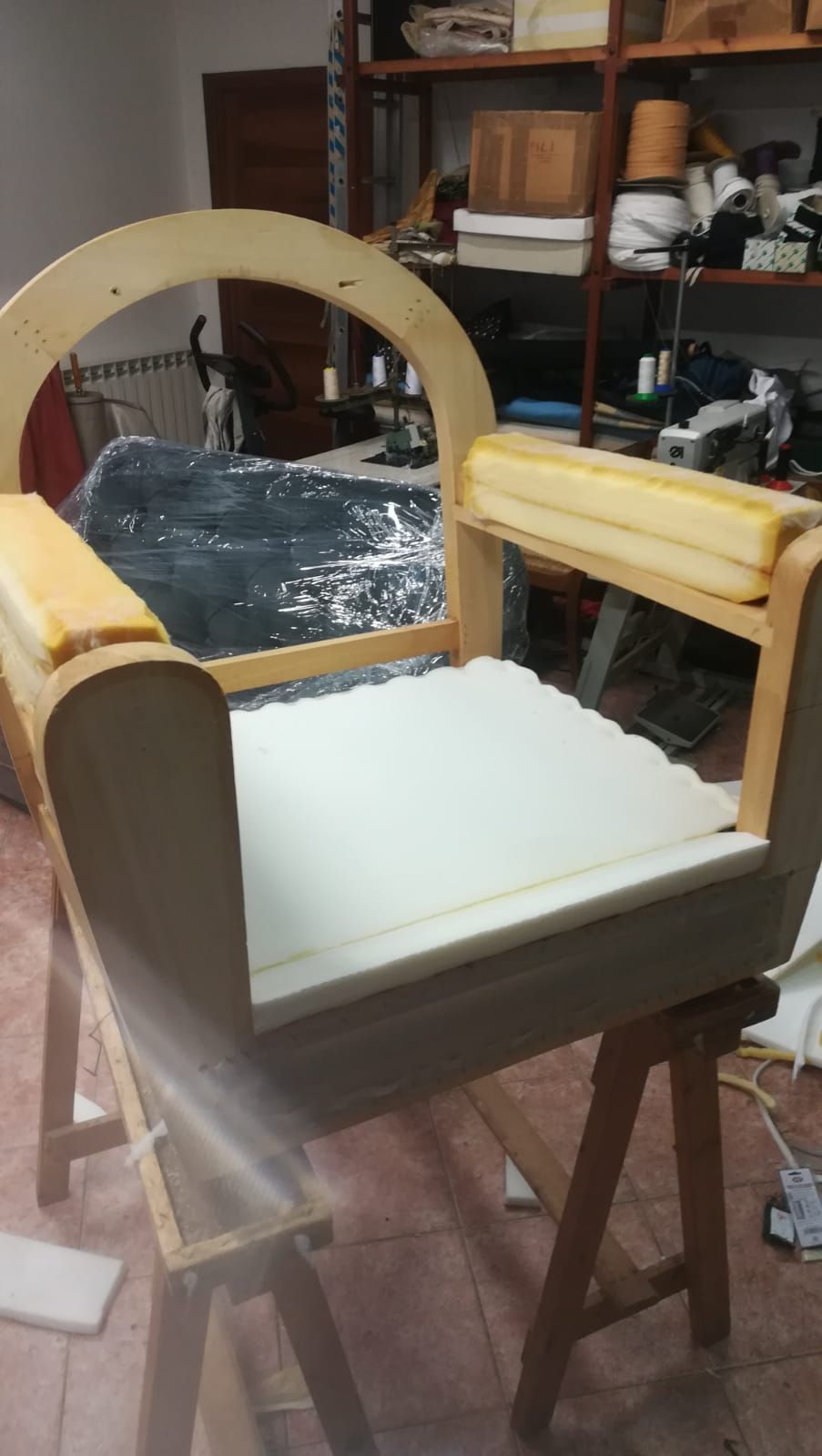 In un'officina, la struttura di una sedia in legno è parzialmente imbottita con cuscini di gommapiuma gialla sui braccioli e gommapiuma bianca sulla seduta.