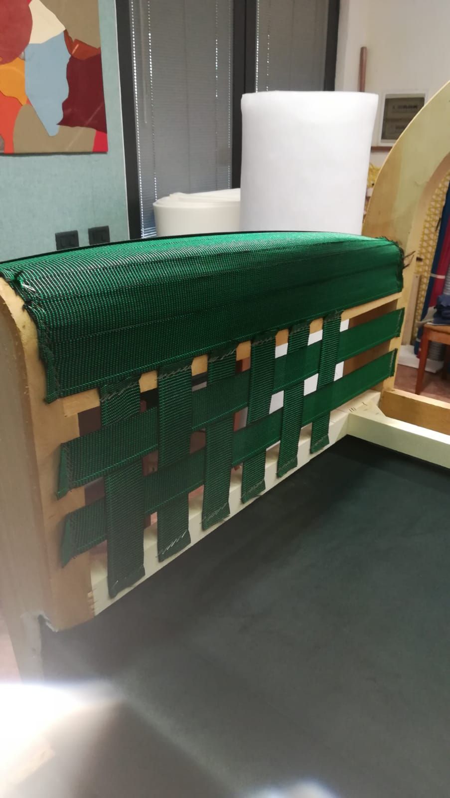 Un tessuto intrecciato di colore verde scuro, fissato a una struttura di legno per la testiera del letto, con un rotolo di tessuto bianco sullo sfondo.