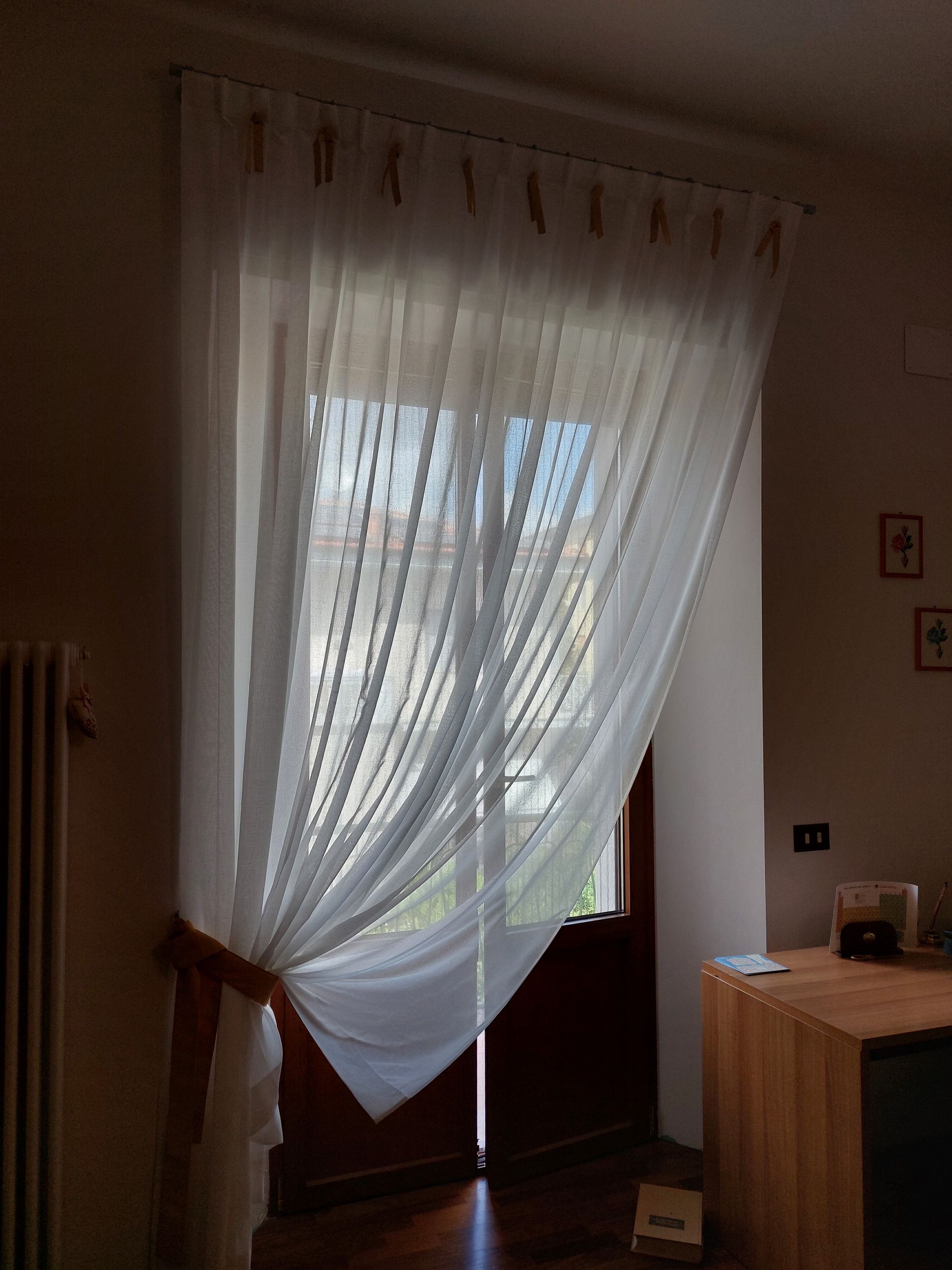 In una stanza illuminata da una luce calda, una tenda bianca e trasparente copre una porta, raccolta da un lato da un fermatenda marrone.