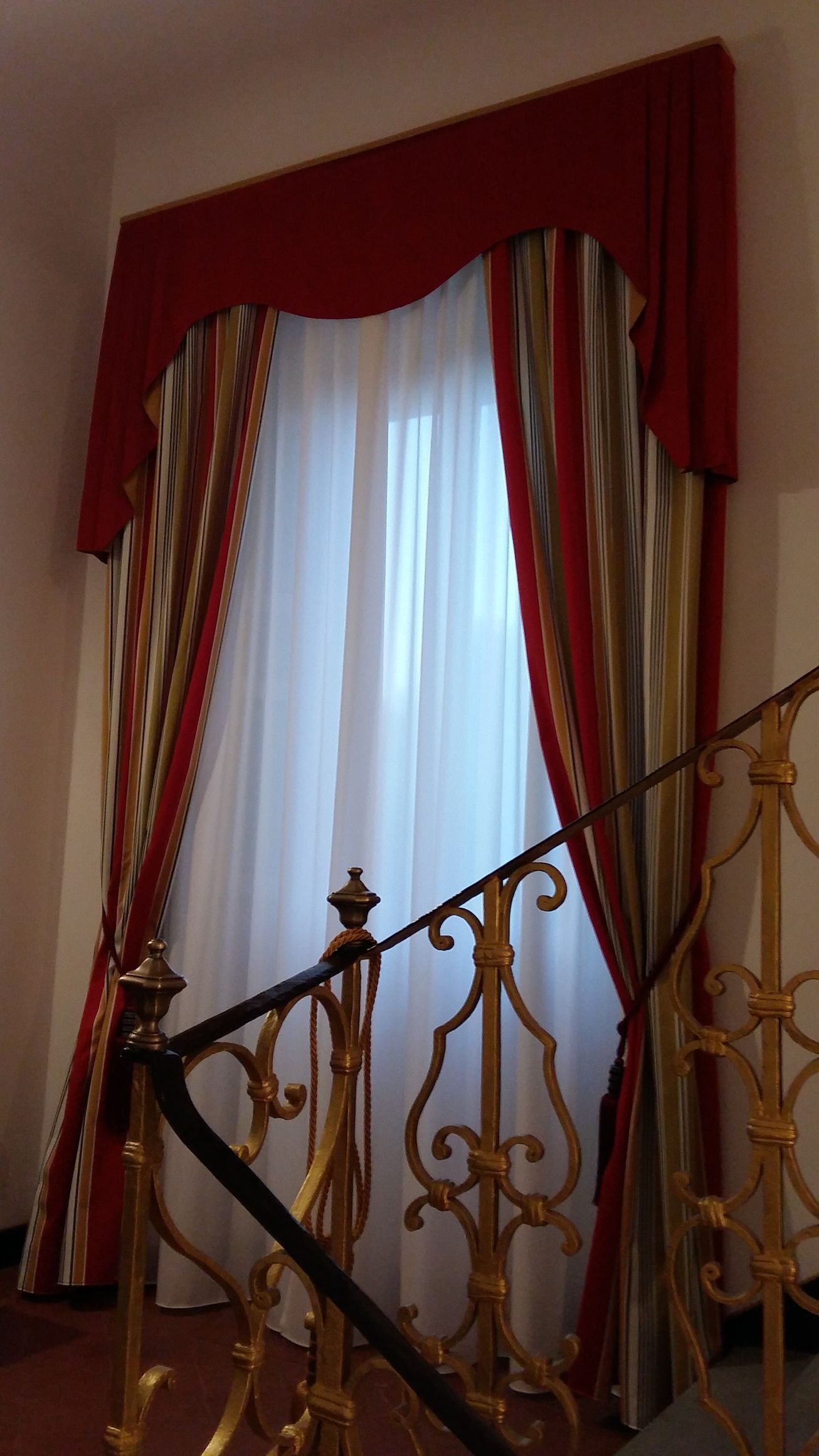 Una finestra con tende bianche trasparenti, drappeggi decorativi rossi e a righe e una mantovana rossa dietro una scalinata dorata riccamente decorata.