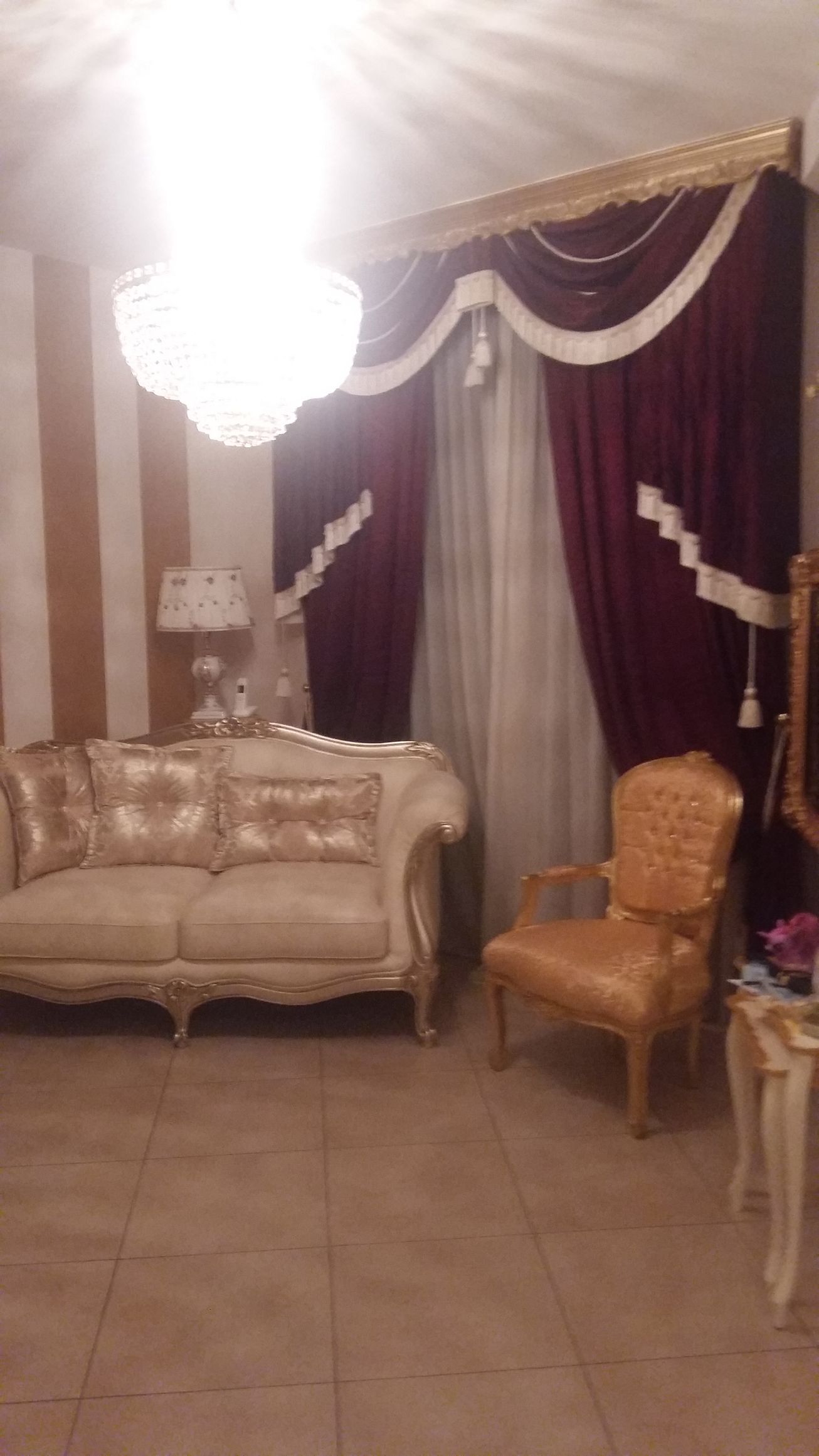 Il soggiorno è arredato con un divano trapuntato beige, una poltrona abbinata, un lampadario di cristallo e tende di velluto rosso scuro.