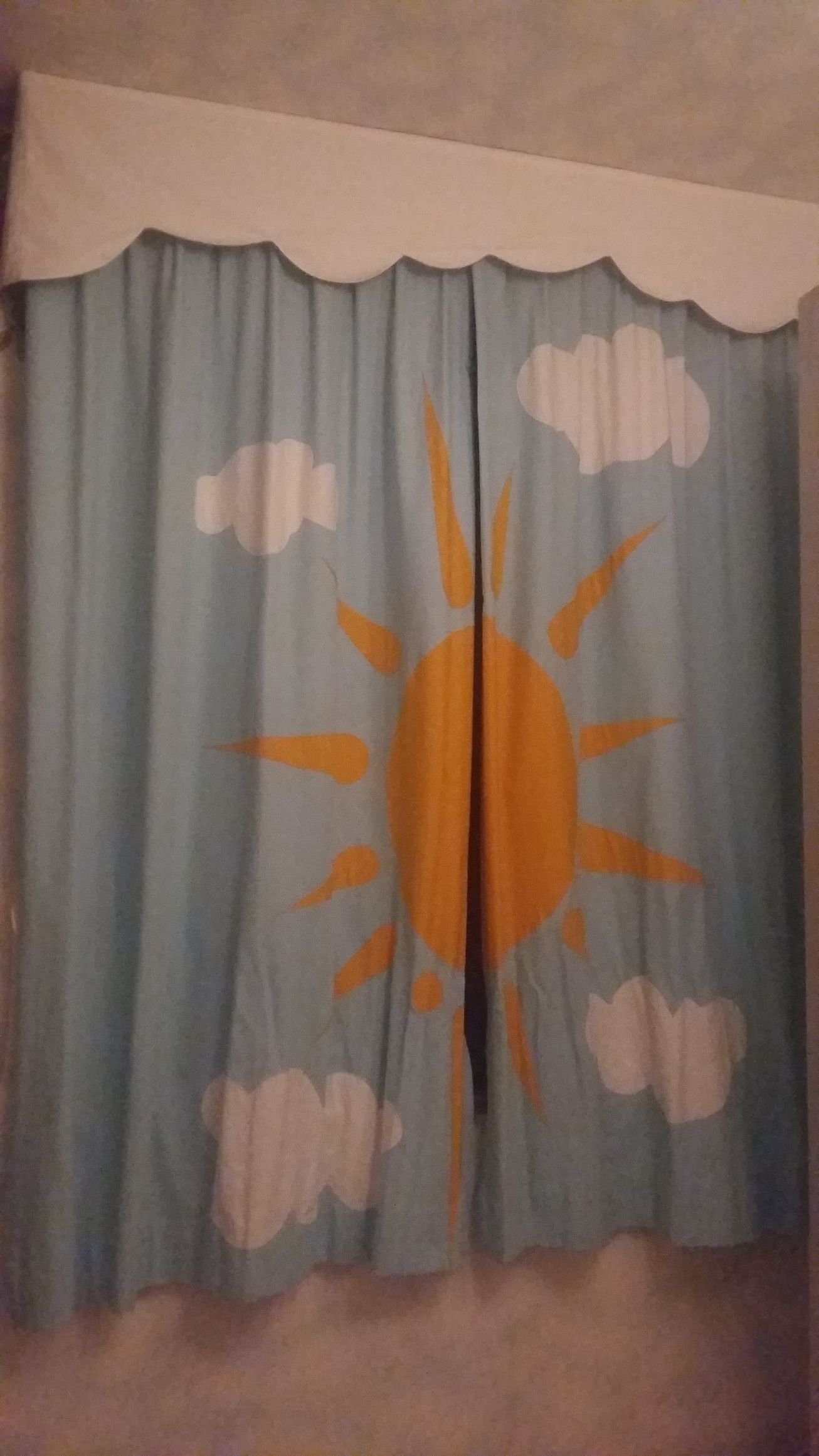 Tende azzurre decorate con un grande sole giallo e nuvole bianche, sormontate da una mantovana bianca smerlata.