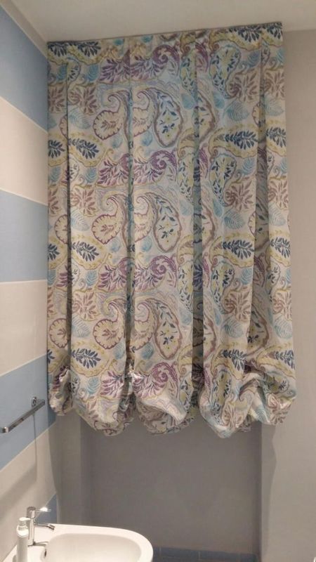 Sopra un lavabo del bagno si trova una finestra coperta da una tenda drappeggiata con motivo paisley nei toni del blu, del viola e del crema.
