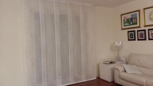 Un soggiorno con tende bianche trasparenti che coprono una finestra, un divano bianco, un tavolino con una lampada e quadri appesi alle pareti.