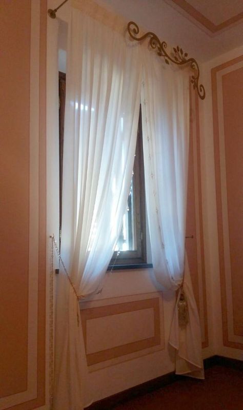 Tende bianche trasparenti pendono da un'asta dorata decorata con motivi a volute, su pareti color pesca con modanature bianche.
