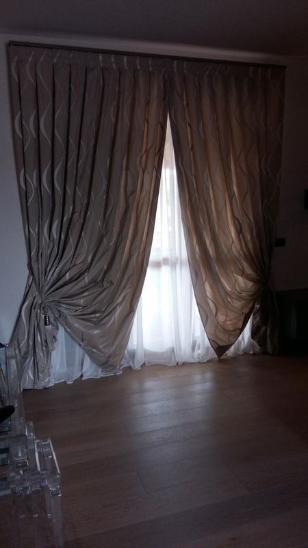 Tende beige a fantasia tirate lateralmente sopra tende bianche trasparenti che coprono una finestra, con pavimento in legno chiaro.