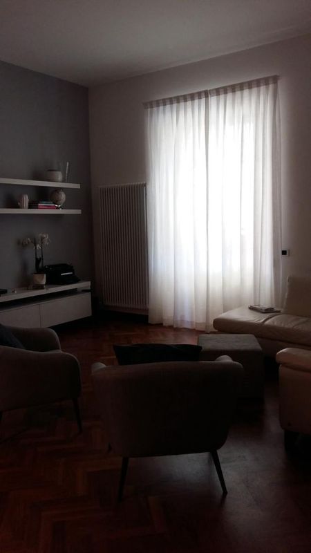 Un soggiorno scarsamente illuminato con pavimento in legno scuro, due poltrone, un divano, un mobile porta TV bianco e tende trasparenti alle finestre.