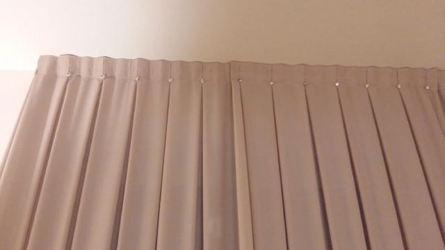 Primo piano di due tende di colore chiaro appese a un binario, caratterizzate da pieghe verticali uniformi.