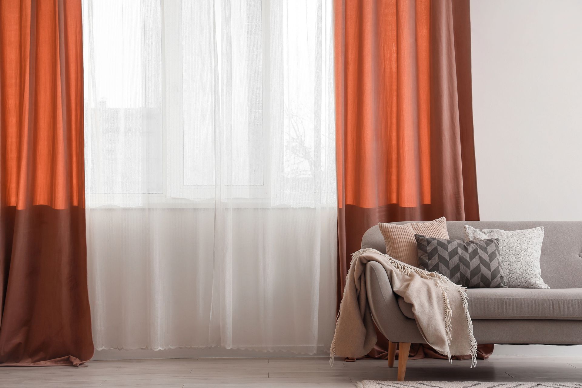 Un divano grigio con cuscini decorativi e una coperta è posizionato davanti a una finestra con tende arancioni e marroni.