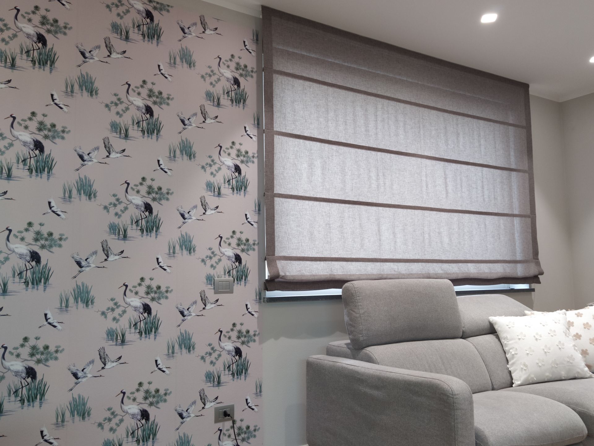 Una stanza con carta da parati a motivi floreali raffiguranti uccelli, una tenda a pacchetto grigia a una finestra e un divano grigio.