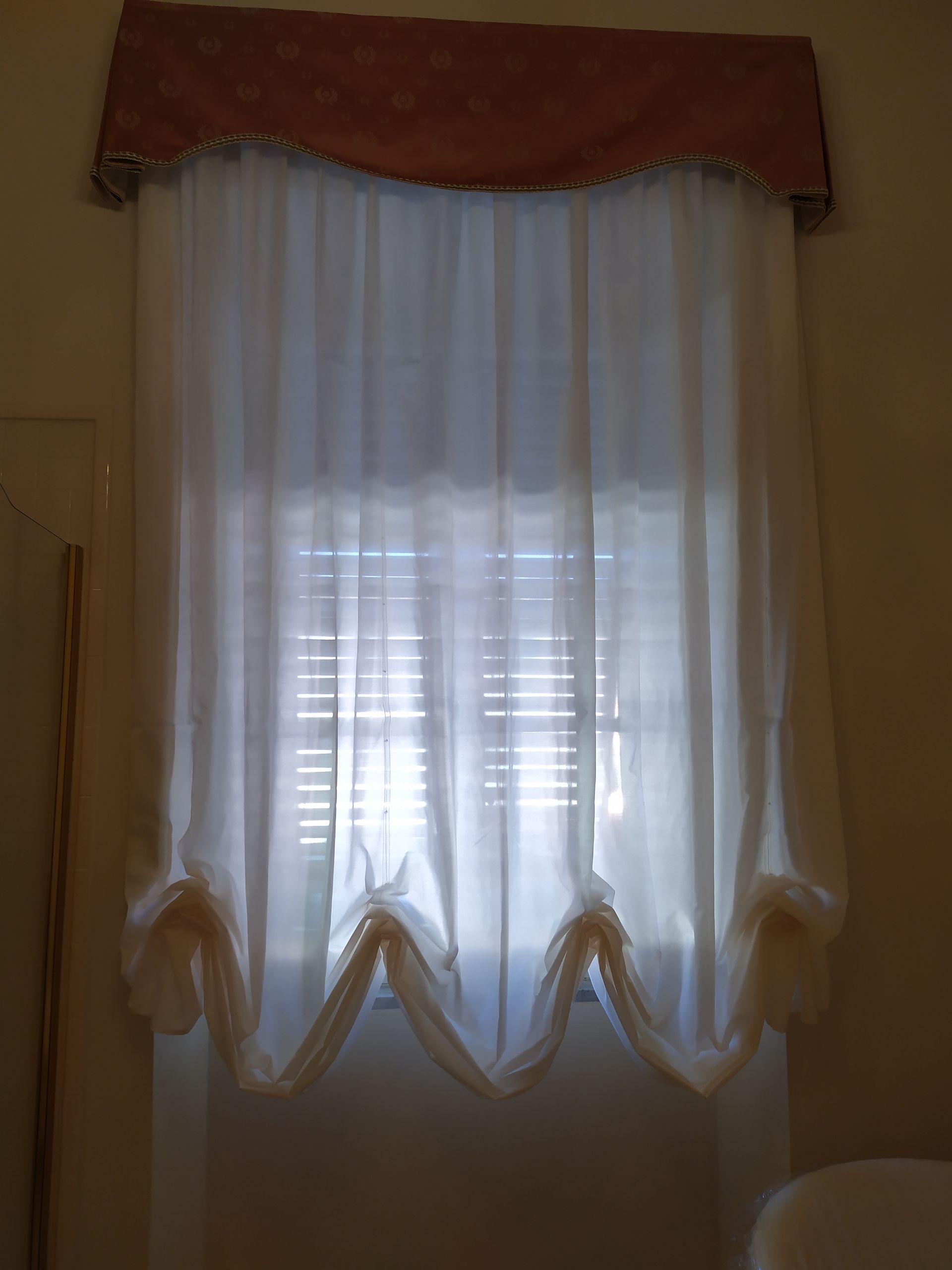 Una tenda bianca arricciata pende da una mantovana decorativa color ruggine sopra una finestra con persiane orizzontali chiuse.
