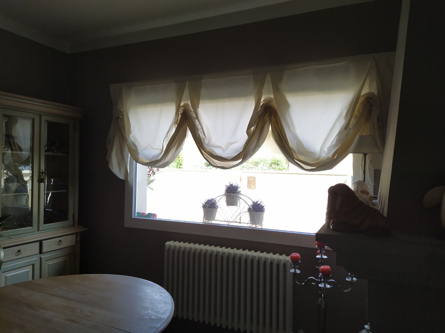 Una sala da pranzo con una finestra con tende trasparenti, un mobile in legno, un tavolo e un termosifone bianco sottostante.