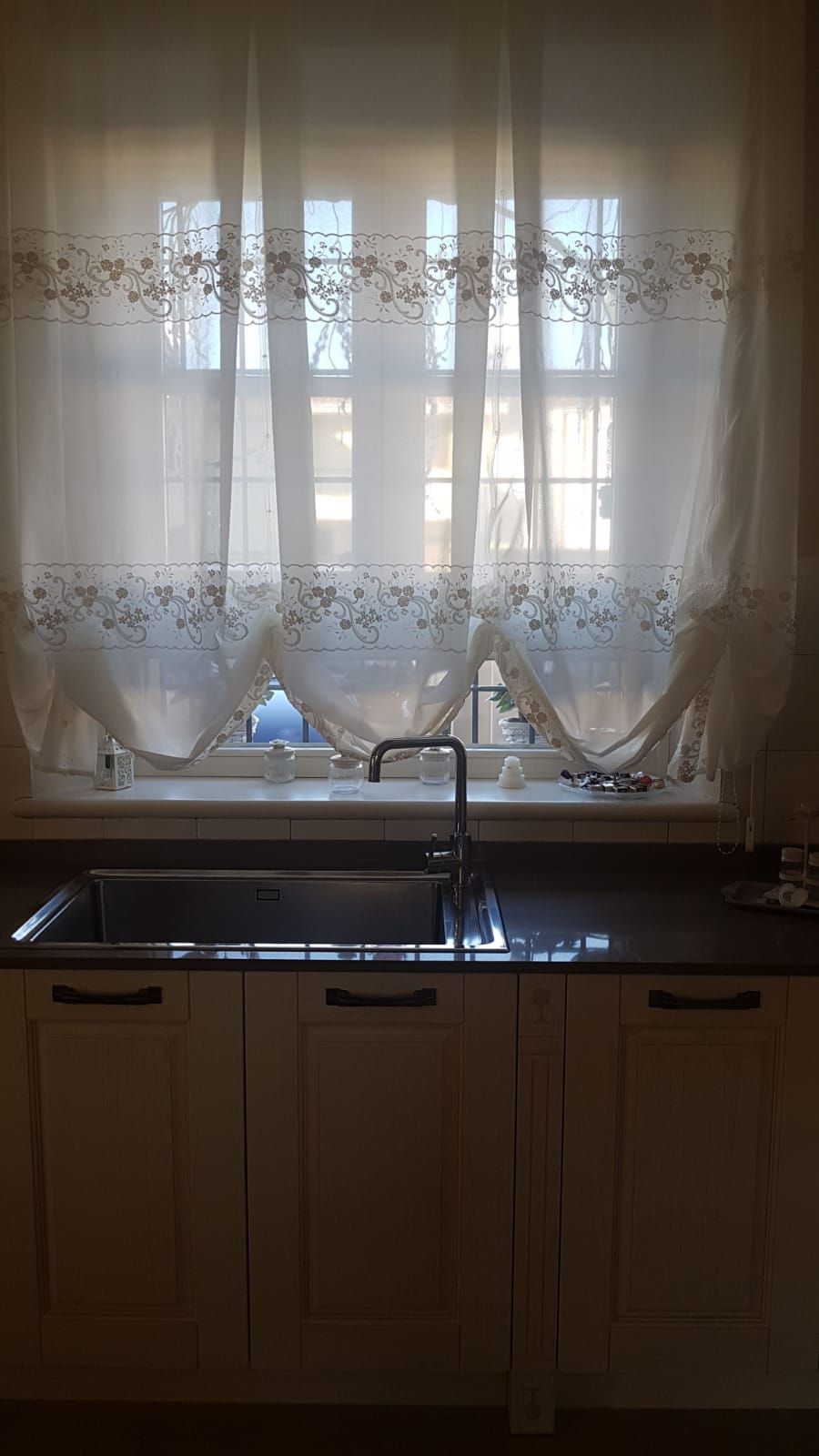 Sotto una finestra coperta da una tenda da caffè ricamata di colore chiaro si trova un lavello da cucina con rubinetto in metallo.