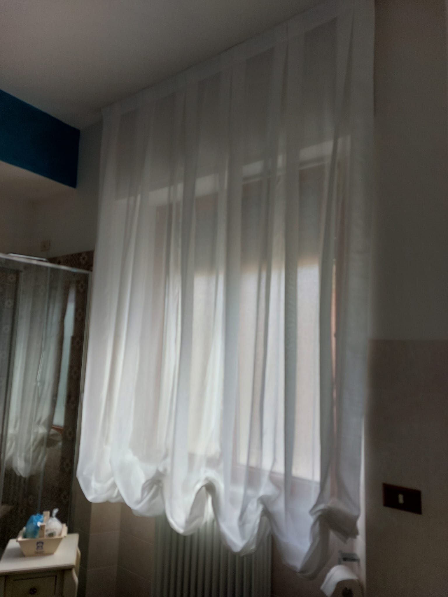 Una tenda bianca trasparente lunga fino al pavimento copre una finestra accanto al mobiletto del bagno e al termosifone.