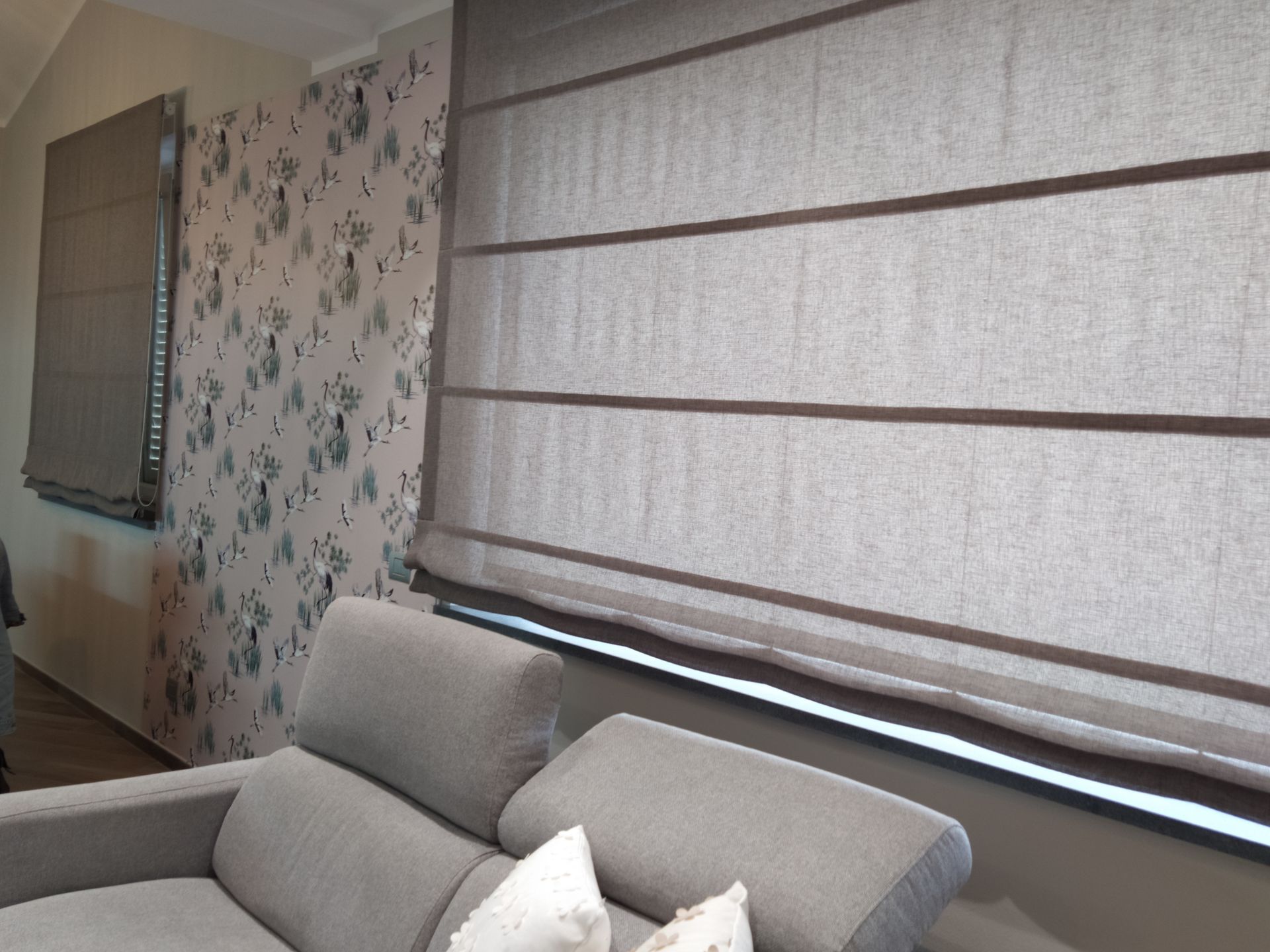 Un divano grigio è posizionato davanti a una finestra con tende a pacchetto marroni, accanto a una parete rivestita di carta da parati a motivi floreali.