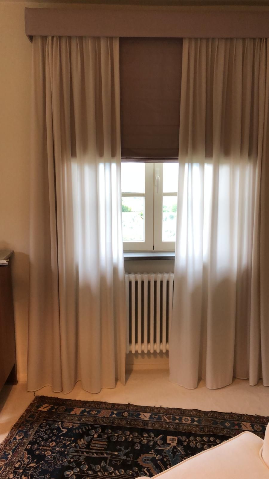 Una finestra con un termosifone sottostante, drappeggiata con tende trasparenti e una mantovana marrone sopra un tappeto a motivi geometrici.