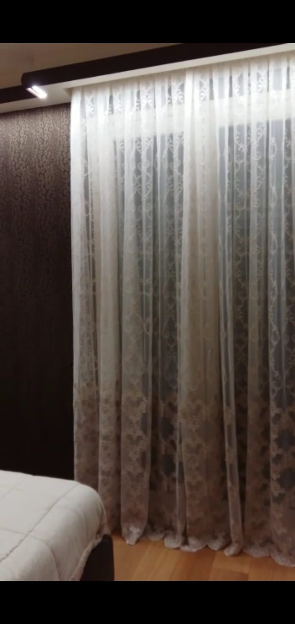 In una camera da letto, accanto al letto, una tenda trasparente a fantasia scorre su un binario contro una parete scura.