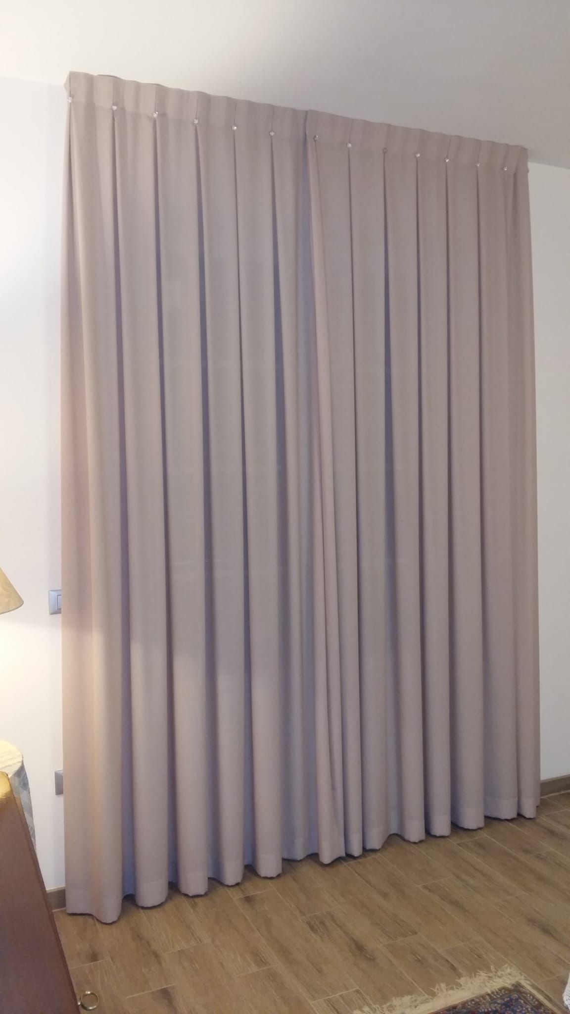 Tende plissettate lunghe fino ai piedi, di colore beige chiaro, appese a un'asta contro una parete bianca sopra un pavimento in legno.