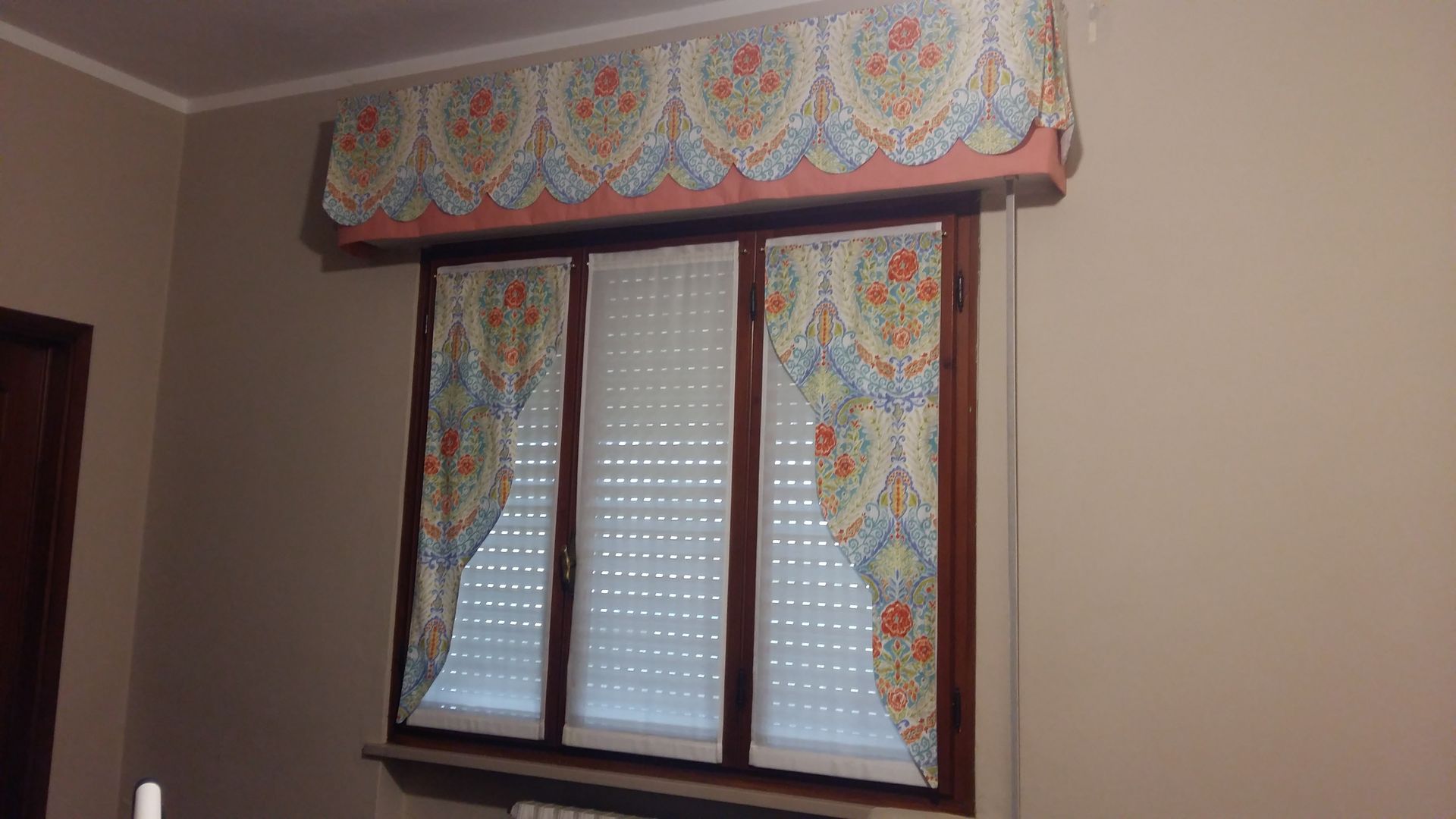 Una finestra con infissi in legno, tapparelle bianche e una mantovana decorativa con motivi floreali e tende laterali abbinate.