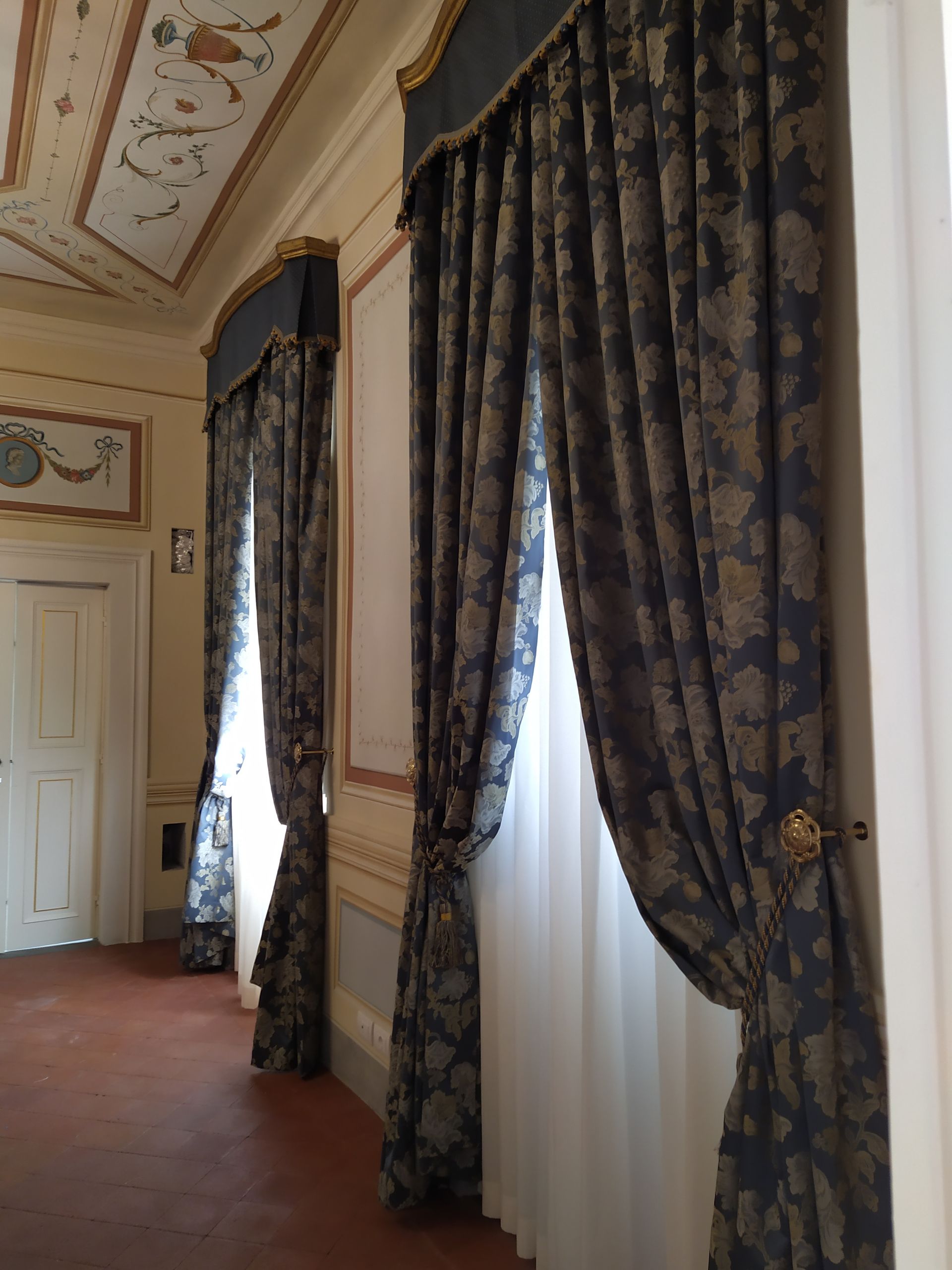 In una stanza riccamente decorata con soffitti affrescati, due alte finestre drappeggiate con tende blu scuro a motivi geometrici e mantovane abbinate.