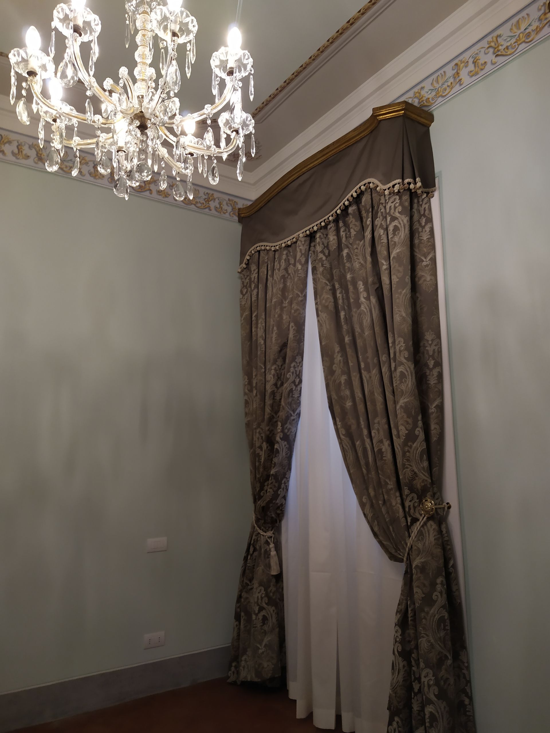 In una stanza dalle pareti azzurre, impreziosita da elaborate modanature e tende lunghe fino al pavimento con motivi decorativi, pende un lampadario di cristallo.