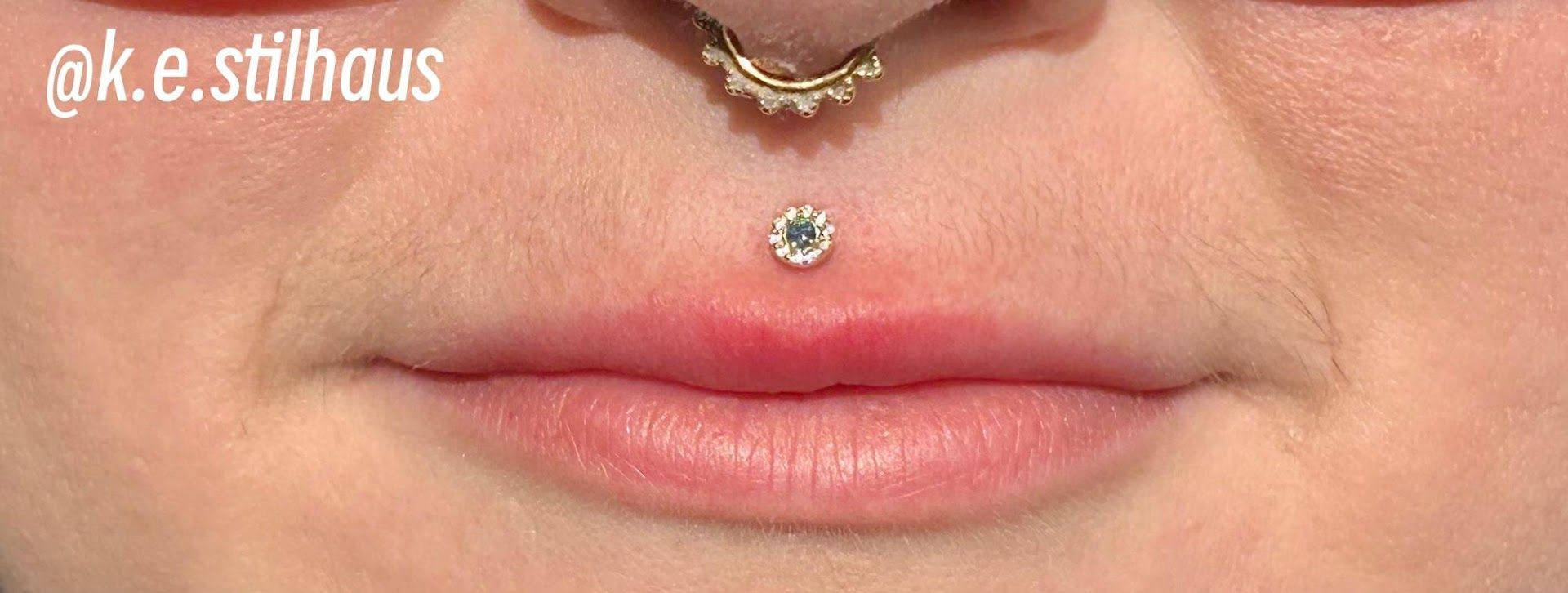 Close-up of a septum piercing with a gold beaded ring and a philtrum piercing with a clear, sparkling gem.