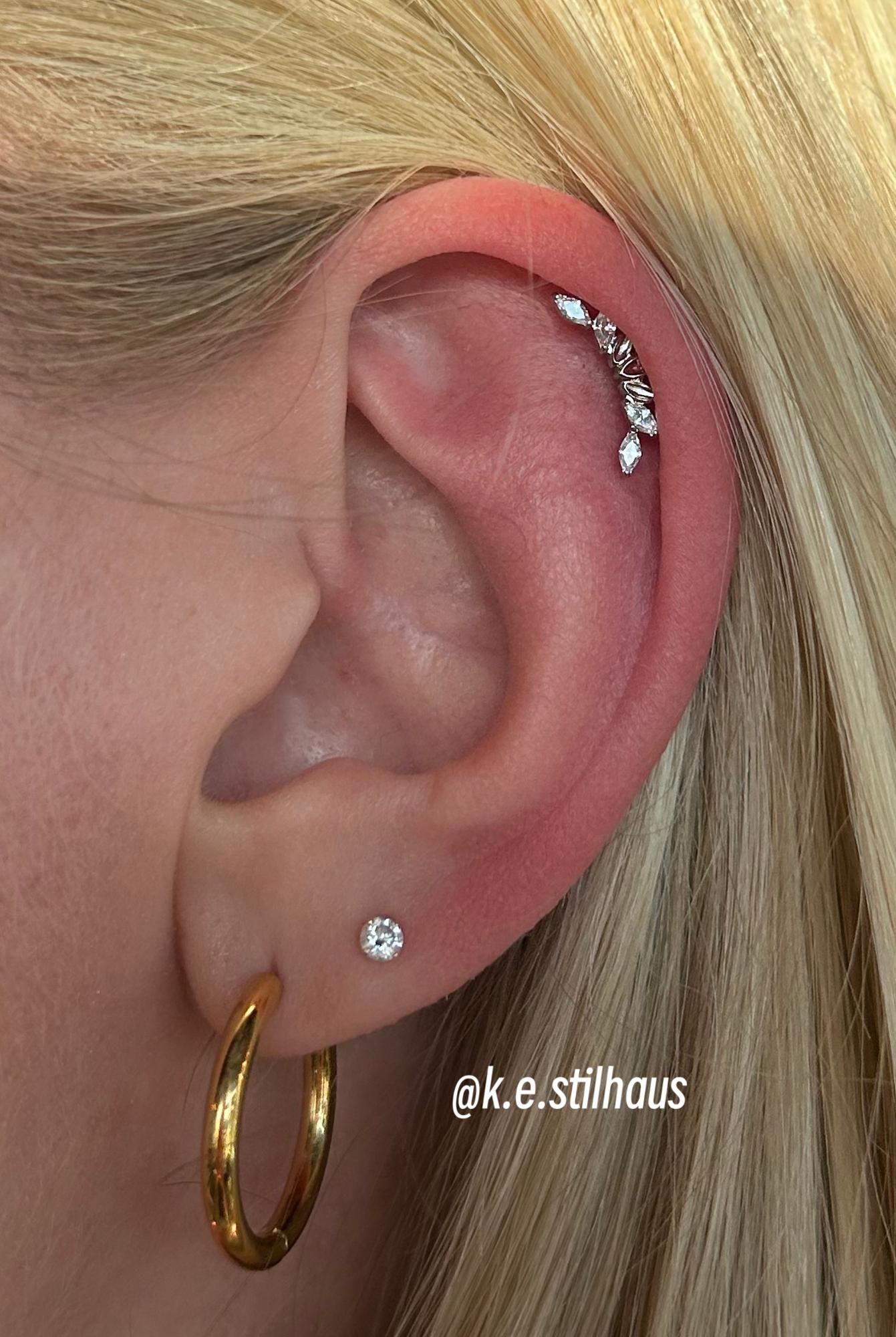 A close-up of an ear featuring a gold hoop, a small stud, and a curved, gem-encrusted climber earring on the upper cartilage.