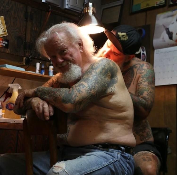 An artist tattooing a person's upper back in a workshop setting with dim lighting and wood-paneled walls.