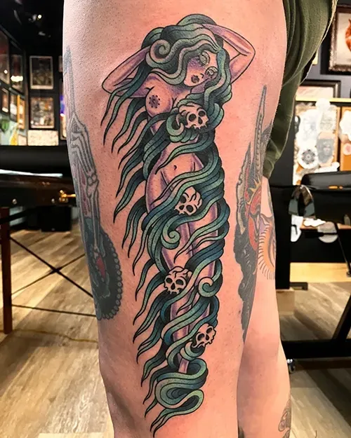 A tattoo on a person's thigh featuring a figure with long, teal, wavy hair intertwined with four small skulls.