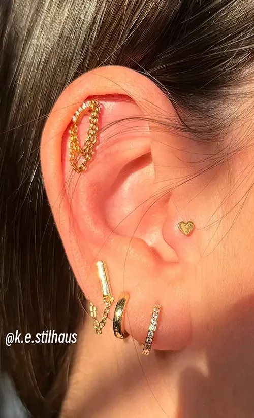 An ear adorned with multiple gold piercings, including a chain cartilage hoop, a heart tragus stud, and three lobe hoops.