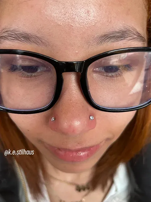 A close-up of a person wearing black-rimmed glasses and two small, sparkling silver stud piercings on their nose.