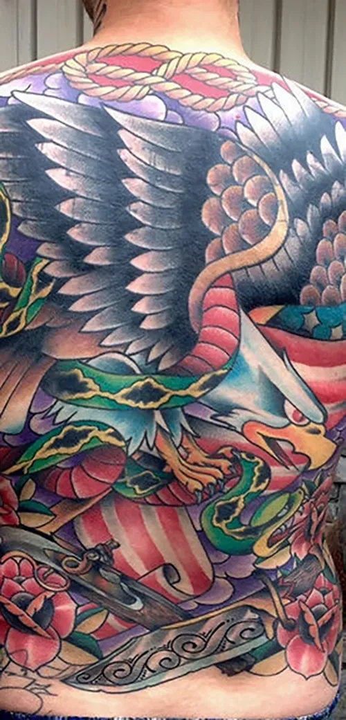 A full-back tattoo featuring a bald eagle fighting a green snake, accented with roses, American flag elements, and rope.