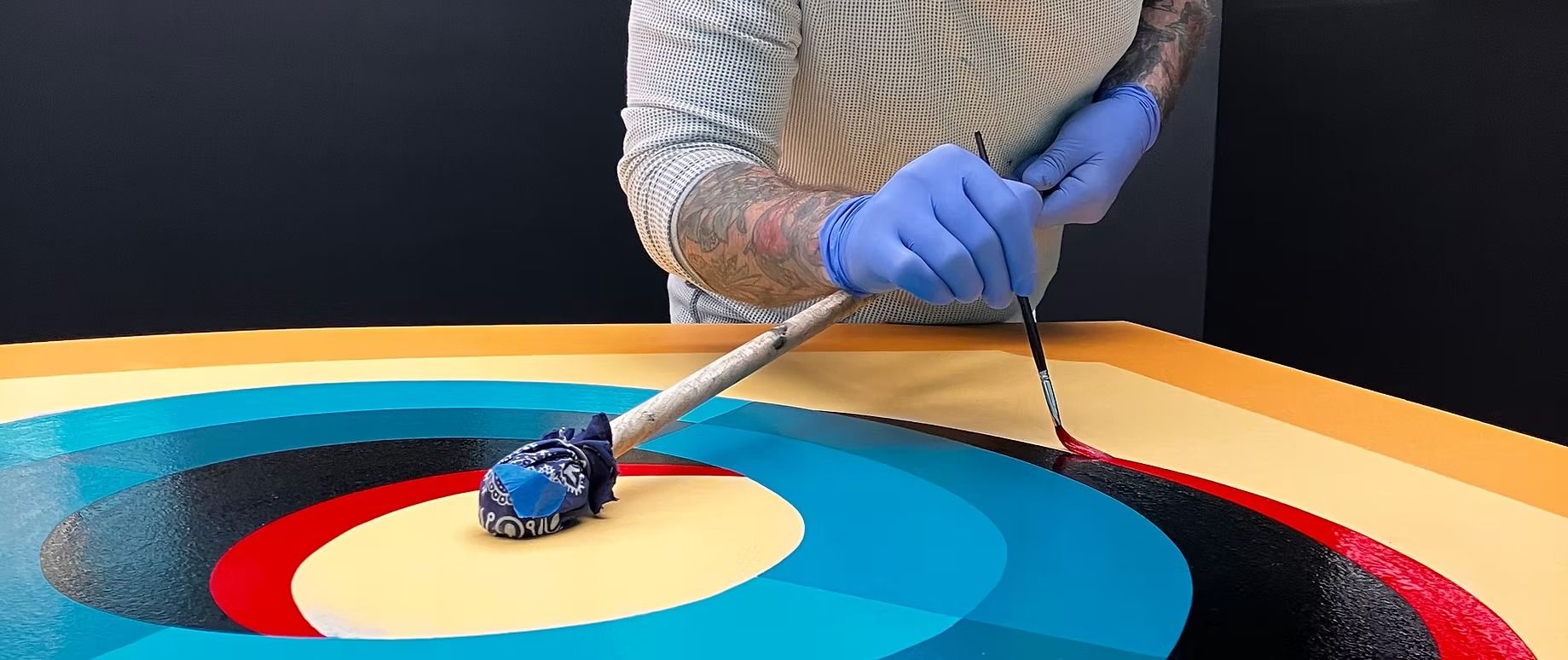 A person wearing gloves uses a brush to paint a pattern of concentric circles in blue, black, red, and yellow on a canvas.