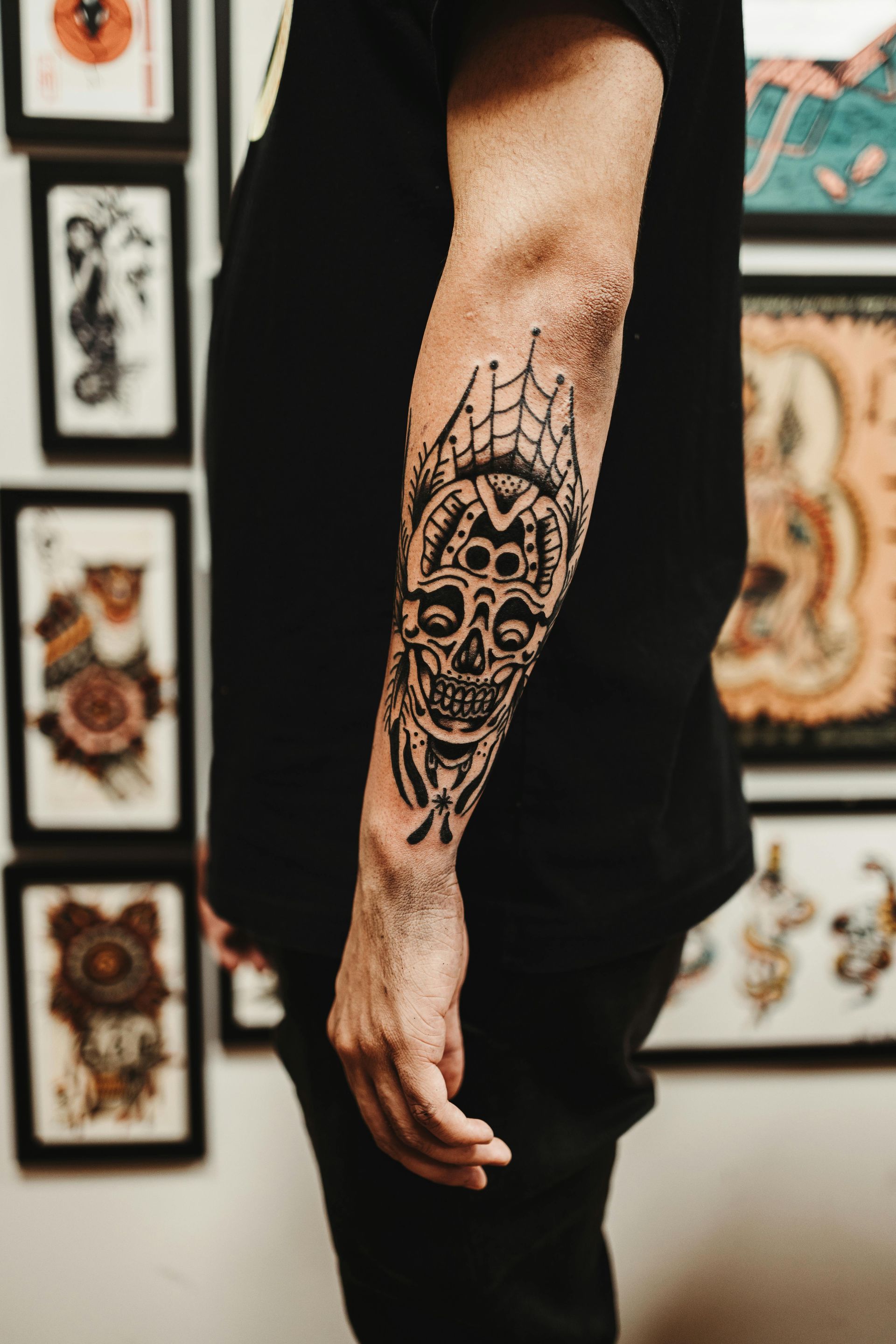 A black ink forearm tattoo featuring a stylized skull and web design, set against a background of framed wall art.