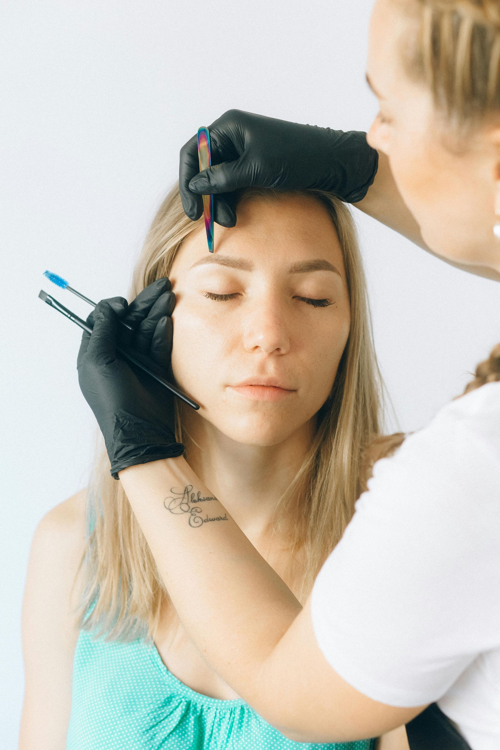 A technician wearing black gloves uses tools to groom the eyebrows of a person with closed eyes in a studio.