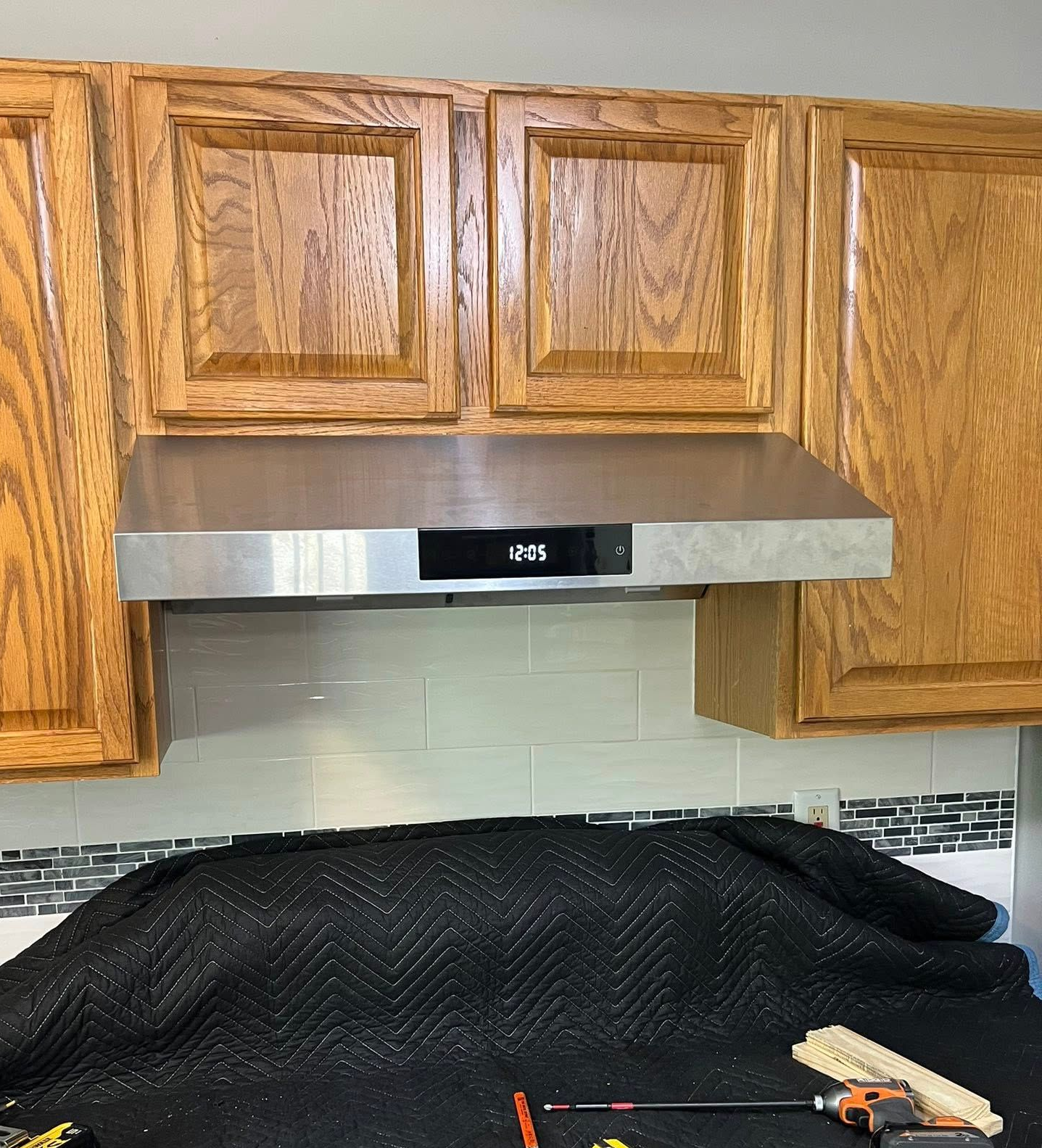 Stainless steel range hood installed under wood kitchen cabinets; backsplash and work in progress covered with a black sheet.