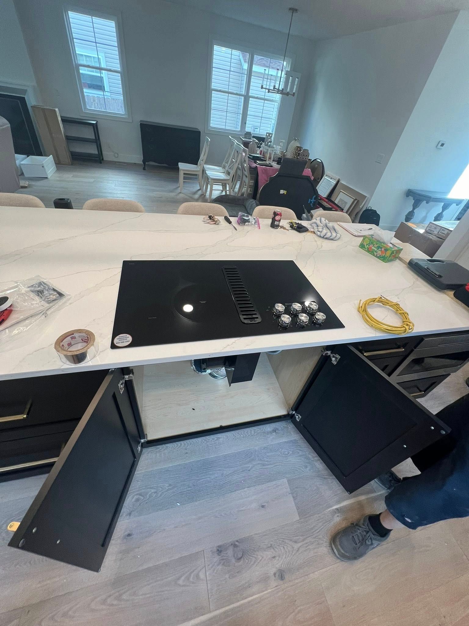 Large kitchen island with open black cabinets, black cooktop, and a person's feet in the corner.