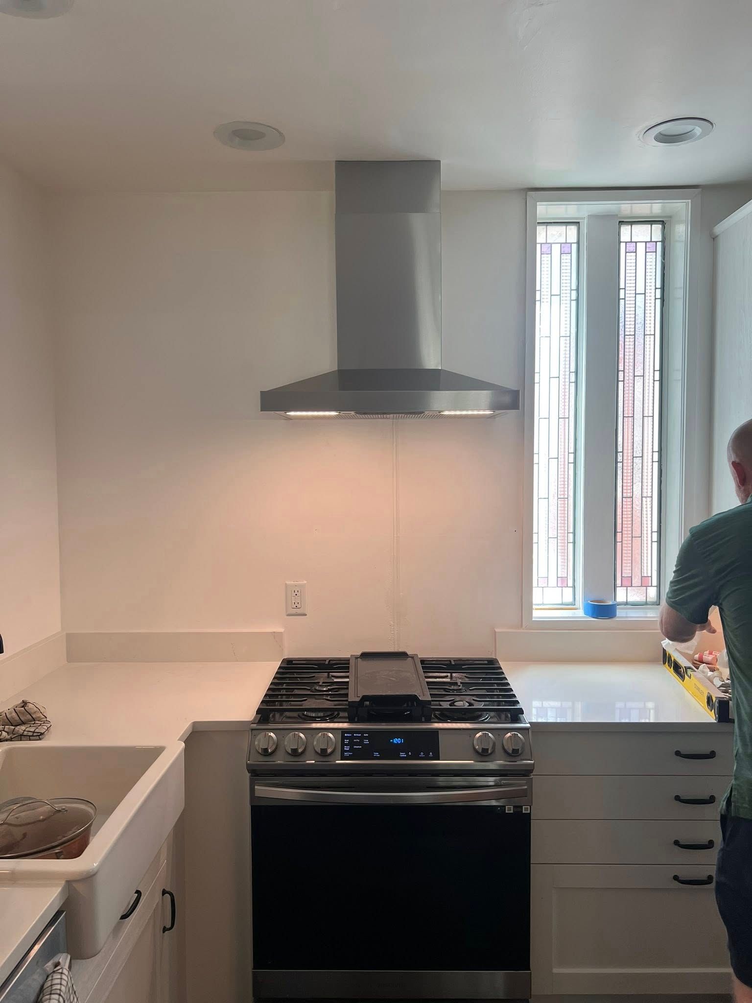 Kitchen with stove, range hood, and a person working near a window. White cabinets and countertop.