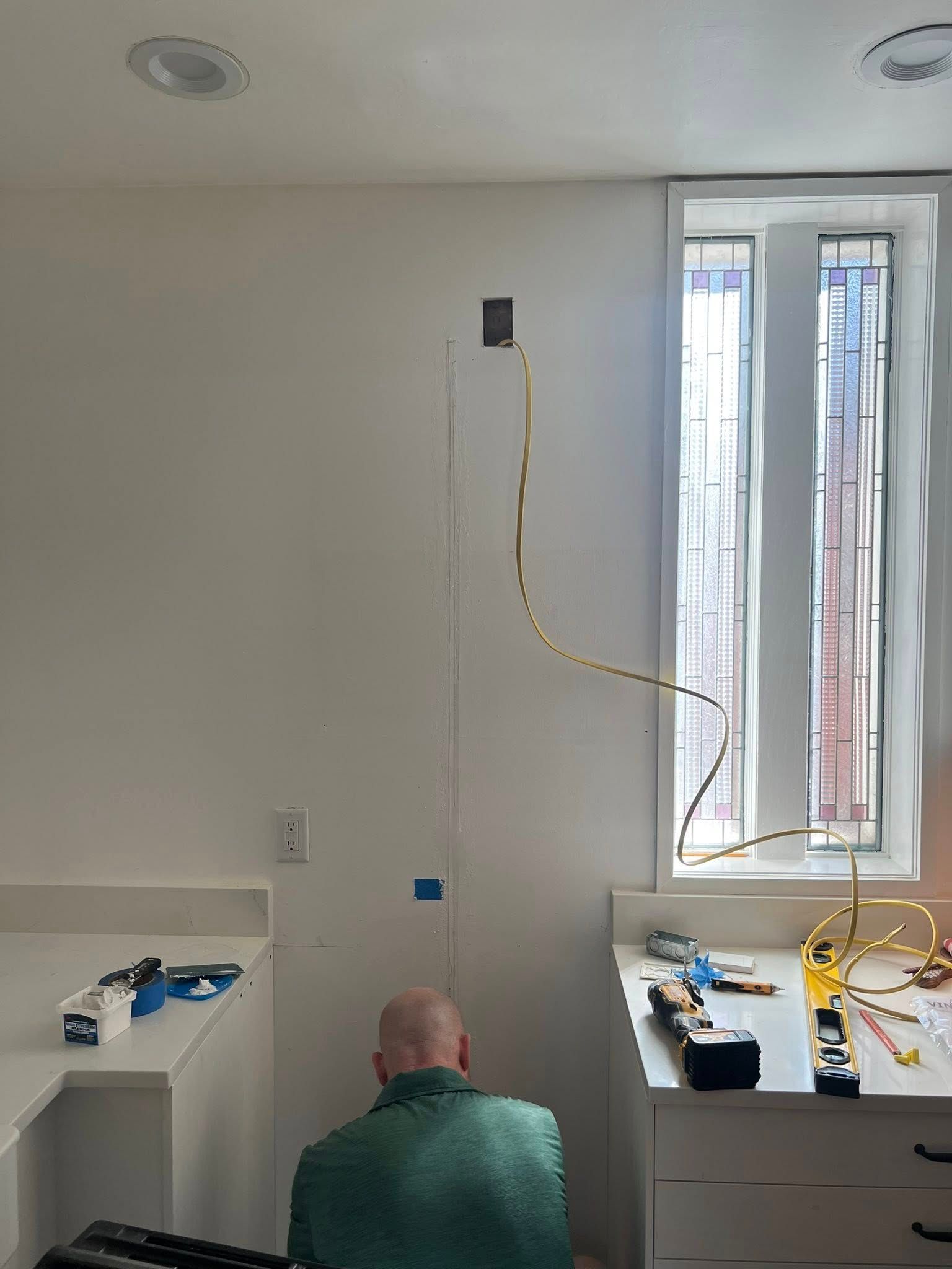 A man working on electrical wiring near a window and white cabinets in a kitchen.