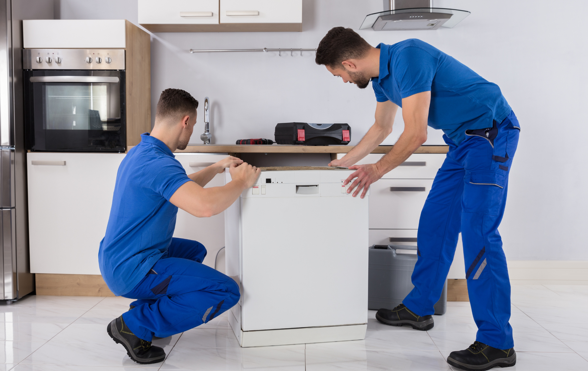 Two men in blue work clothes installing a dishwasher in a kitchen.