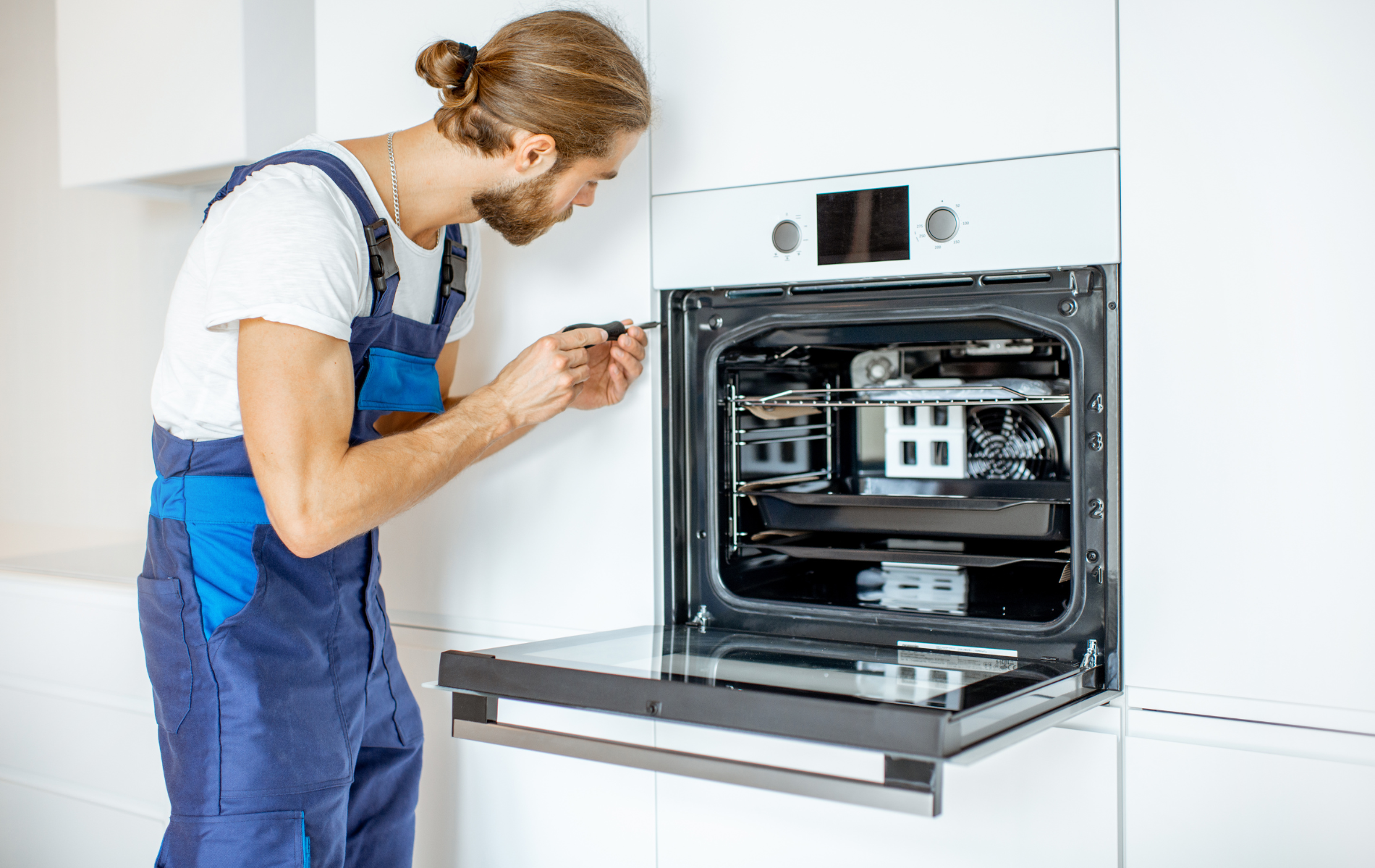 Man in blue overalls repairing an oven in a white kitchen.