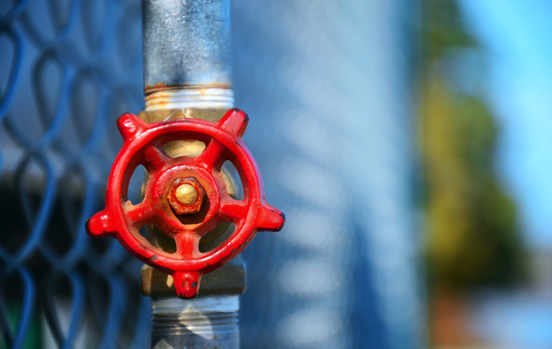 Red valve on a silver pipe against a blurred fence and blue sky.