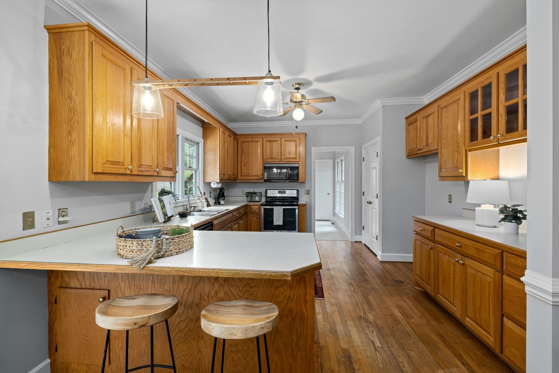 A kitchen with light oak cabinets, white countertops, two wooden bar stools, and hardwood flooring.