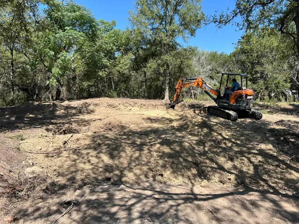 A man is driving an excavator in a dirt field surrounded by trees.