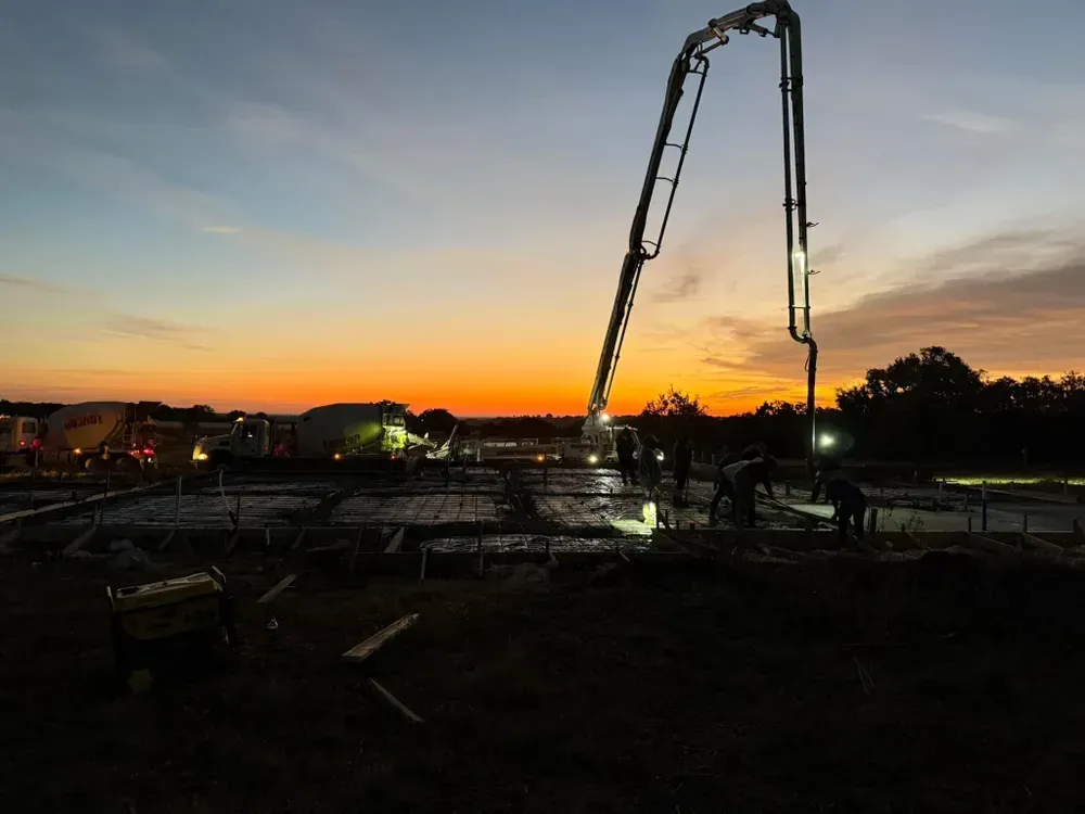 A concrete pump is being used to pour concrete at a construction site at sunset.