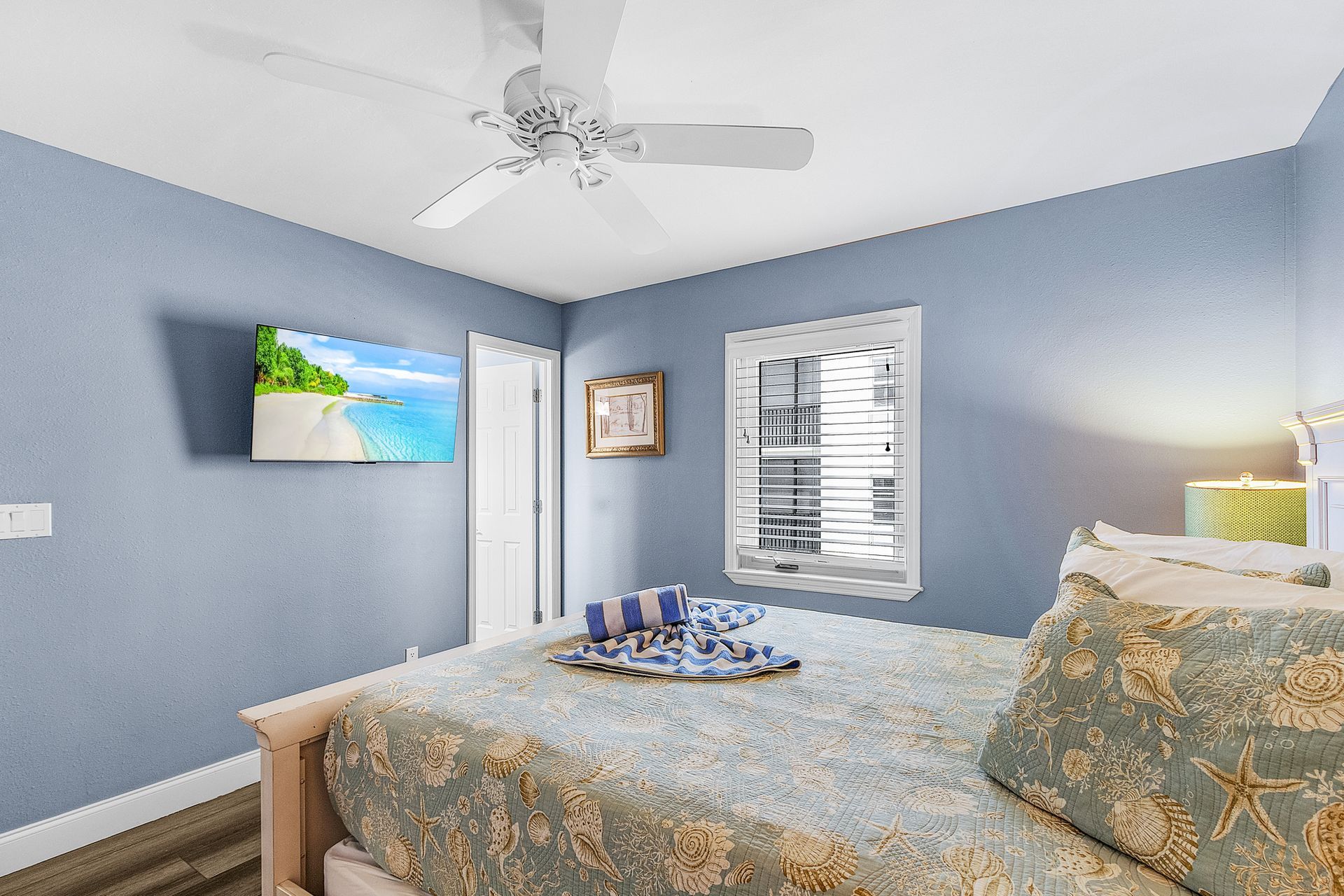 Blue bedroom with a bed, ceiling fan, window blinds, wall TV, and ocean-themed decor.